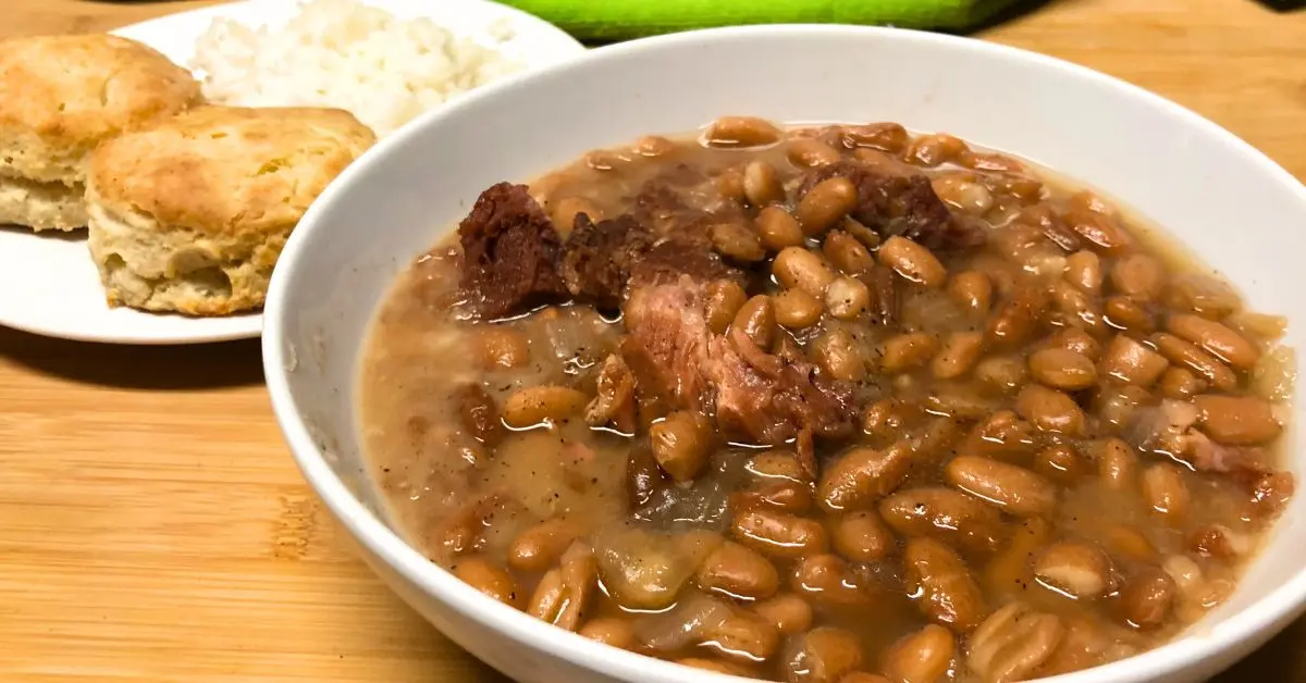 Old-fashioned Southern pinto beans with chunks of smoked meat in a white bowl, served with two golden buttery biscuits and a small mound of white rice on the side.