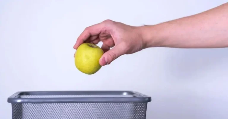 A person discarding a spoiled apple into a silver trash can.