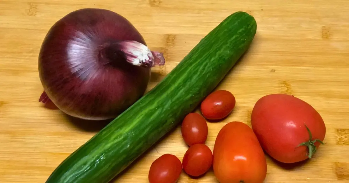Whole cucumber, Roma tomatoes, and red onion on a cutting board for an Israeli salad recipe.