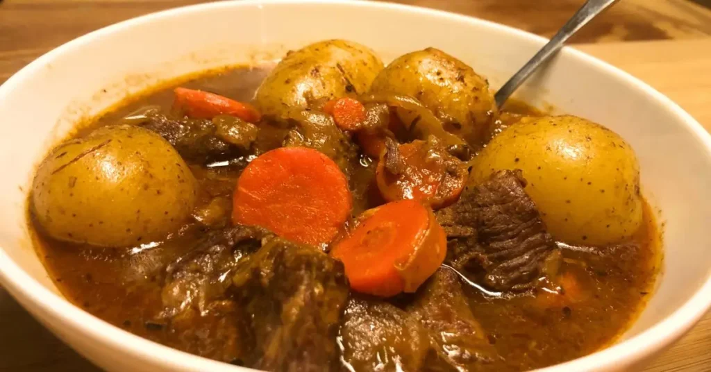 Hearty beef stew without wine in a white bowl with spoon inside with large chunks of chuck roast, whole baby gold potatoes, sliced carrots, and flavorful broth.