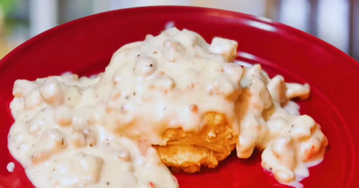A fluffy butter biscuit topped with a generous serving of sausage and gravy, with gravy dripping over the biscuit, served on a red plate.