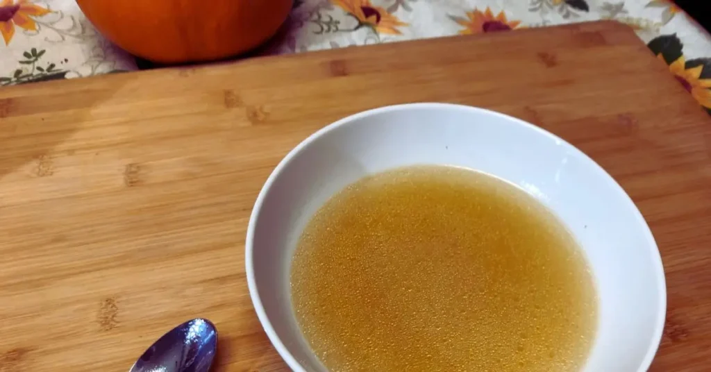 white bowl, filled with golden chicken broth on a wooden board and with spoon and fall decor in the background.