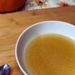 white bowl, filled with golden chicken broth on a wooden board and with spoon and fall decor in the background.