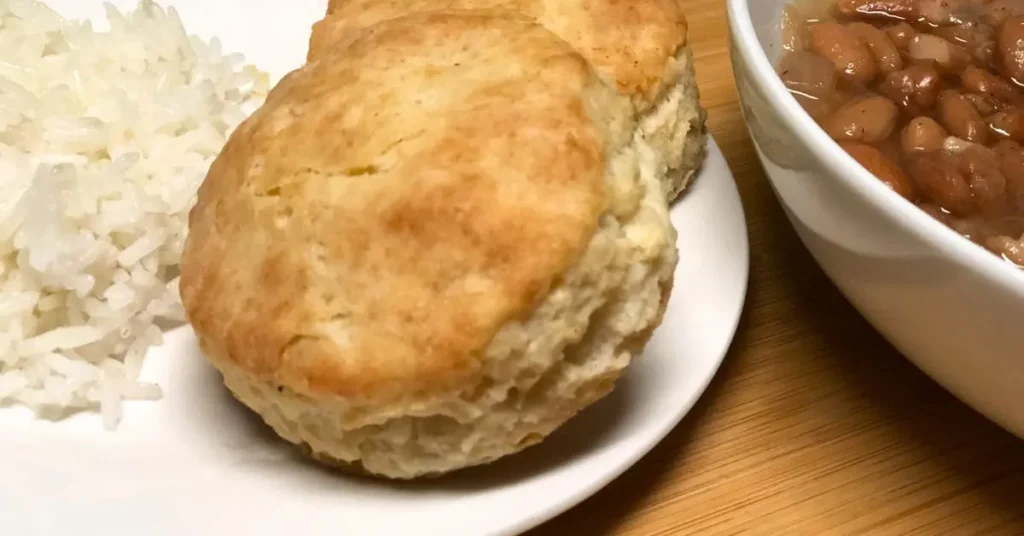 Buttery golden biscuit on a white plate, served with a side of white rice next to a bowl of pinto beans.”
