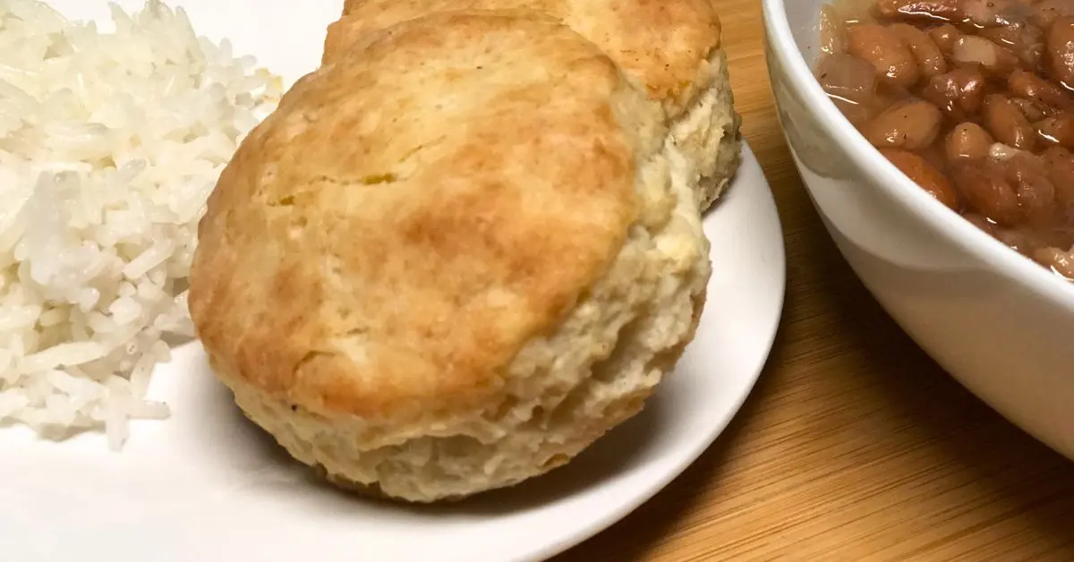 Buttery golden biscuit on a white plate, served with a side of white rice next to a bowl of pinto beans.”