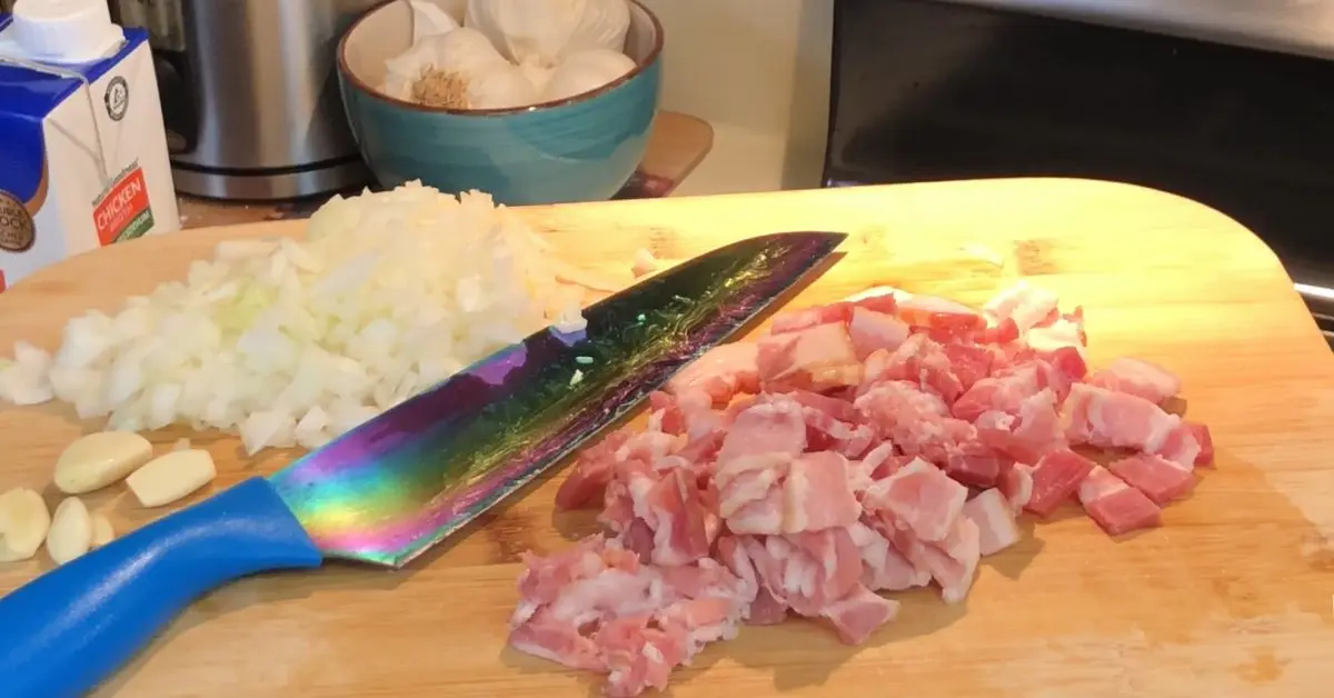 Raw diced bacon and onions on a cutting board with cloves of garlic and a bowl of broth in the background for creamy potato soup