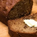 Moist all butter banana bread with one slice cut and topped with butter, served with a cup of tea