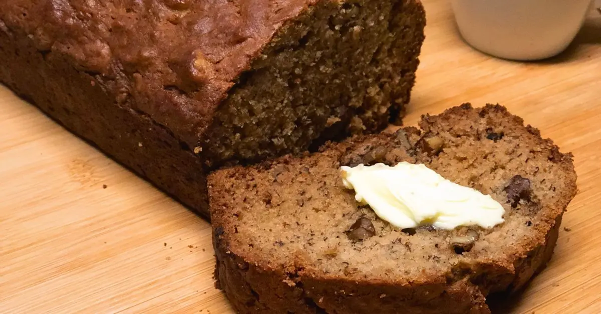 Moist all butter banana bread with one slice cut and topped with butter, served with a cup of tea