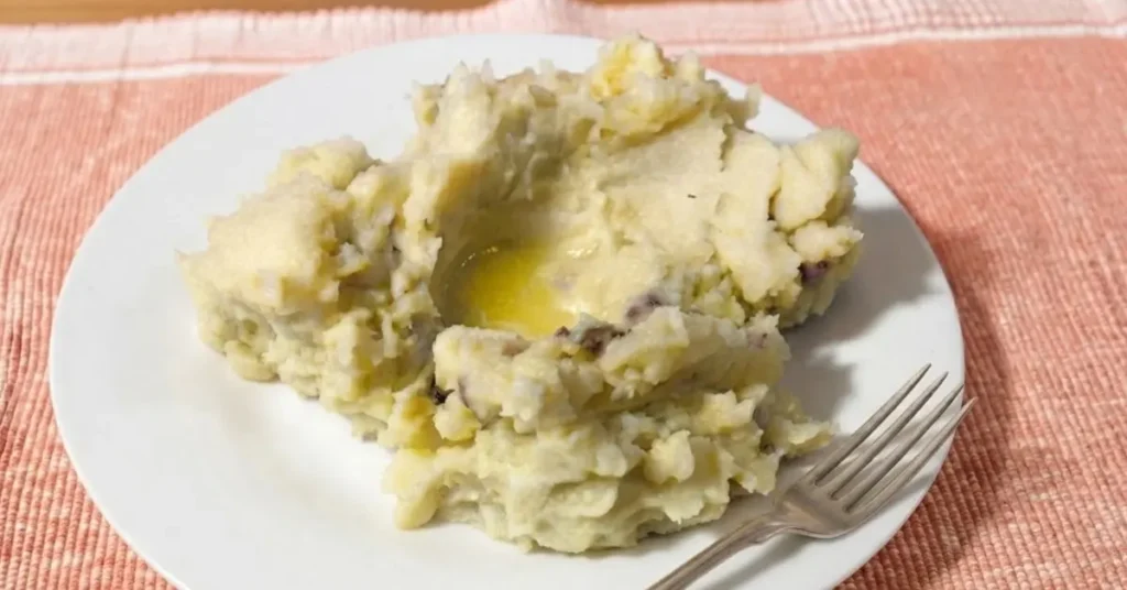 Rustic red skinned mashed potatoes with creamy texture and potato skin on white plate with silver folk on a peach placemat