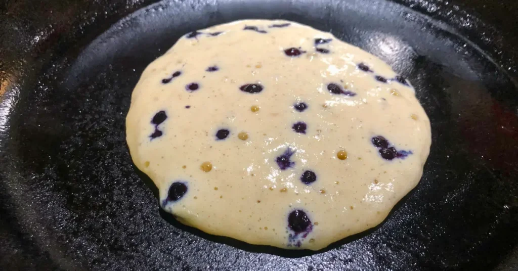 Blueberry buttermilk pancake cooking in a cast iron skillet before being flipped with fresh blueberries on top of the batter.