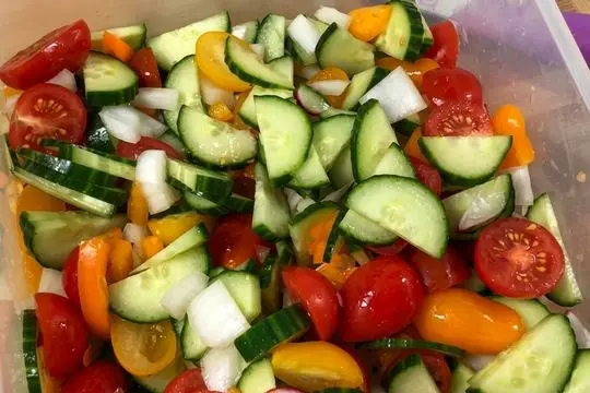 Top-down view of a container of chopped vegetables, including crisp green cucumbers, red and yellow tomatoes, and white onions.
