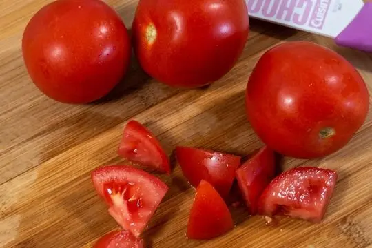 Fresh whole red tomatoes and chopped tomato wedges on a wooden cutting board next to a white and purple knife.