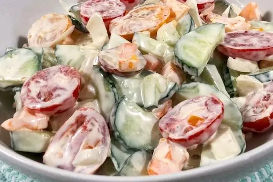 A vibrant bowl of creamy cucumber and tomato garden salad coated in a rich dressing, showing bright red tomato halves and green cucumber slices.