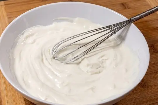 A side angle view of a white ceramic bowl containing a smooth, creamy white salad dressing with a wire whisk resting inside.