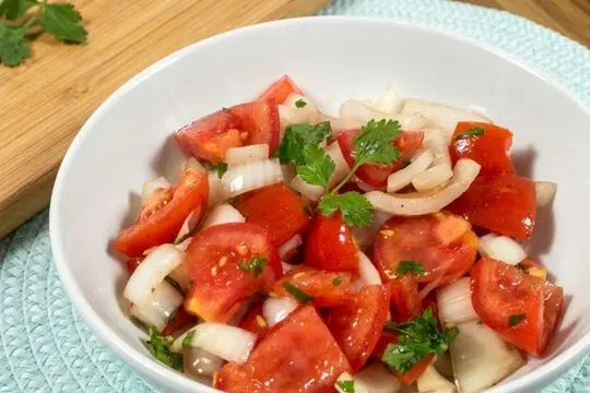 A white ceramic bowl filled with a fresh tomato and onion salad tossed with lemon juice and herbs, resting on a textured light blue placemat.