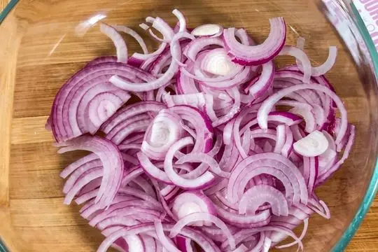 A glass mixing bowl filled with a large batch of thinly sliced red onion rings, ready to be pickled.