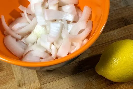 Sliced white onions inside a bright orange mixing bowl sitting next to a whole fresh yellow lemon on a wooden cutting board.