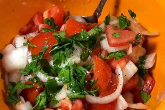Chopped red tomatoes and sliced white onions in an orange bowl topped with freshly chopped green herbs and black pepper before mixing.