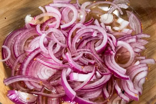 Thinly sliced red onion rings completely submerged and soaking in a clear vinegar brine with mustard seeds inside a clear glass bowl.
