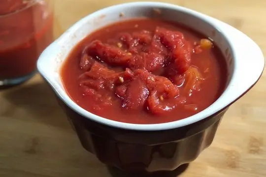 Whole peeled canned tomatoes in a red bowl with reserved tomato juice measured for tomato pudding