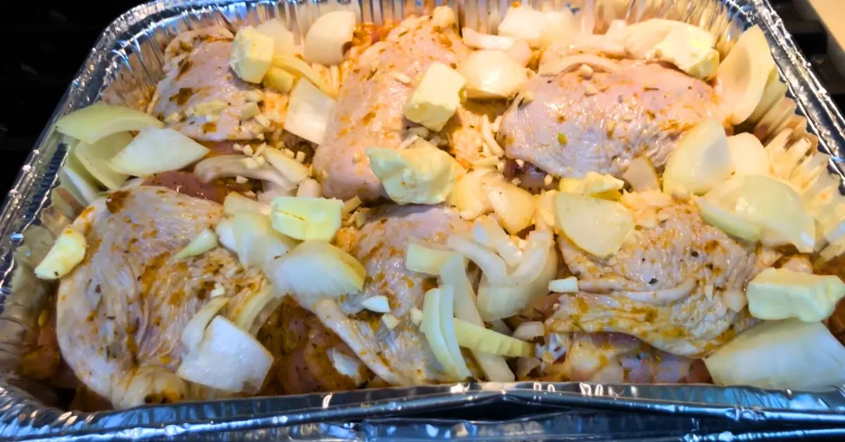 Seasoned chicken thighs arranged in a foil pan with sliced onions and pats of butter.