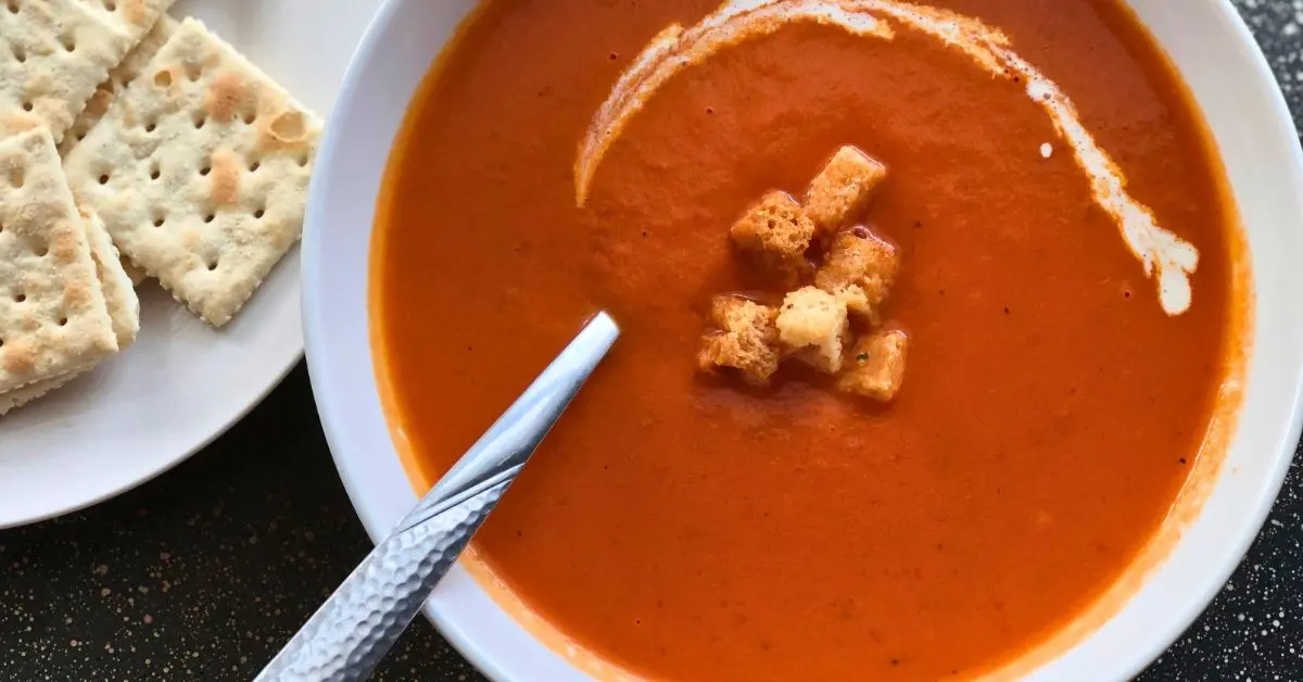 Creamy tomato soup with Parmesan topped with a swirl of cream and croutons in a white bowl with saltine crackers