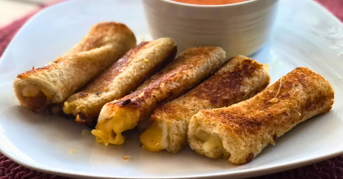 four golden brown garlic bread grilled cheese rollups on a white square plate and a white ramekin of tomato soup dip