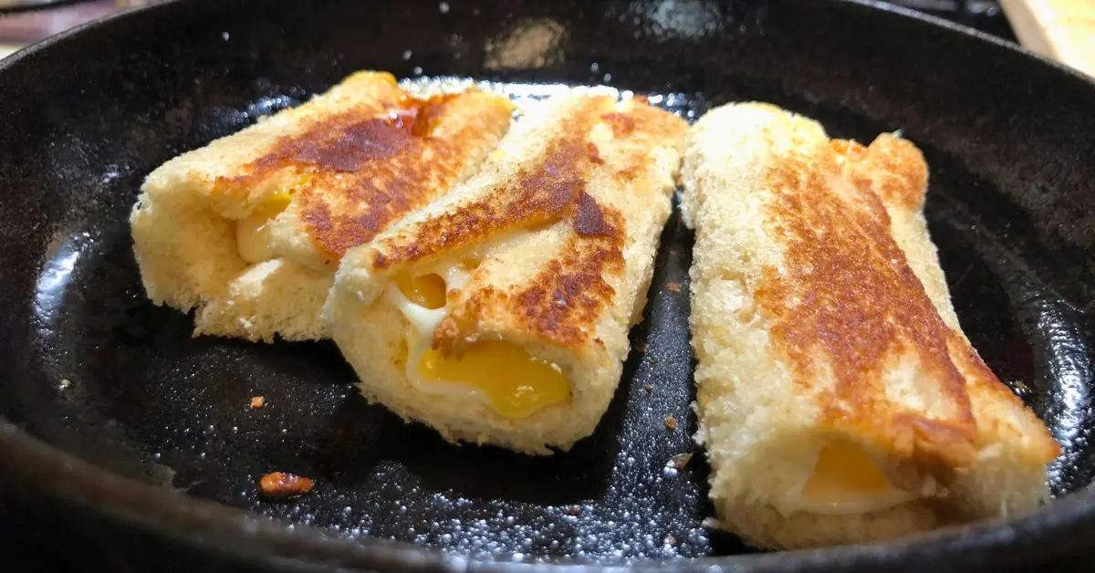 three golden brown grilled cheese rollups in small black iron skillet with melted cheese viewed from side